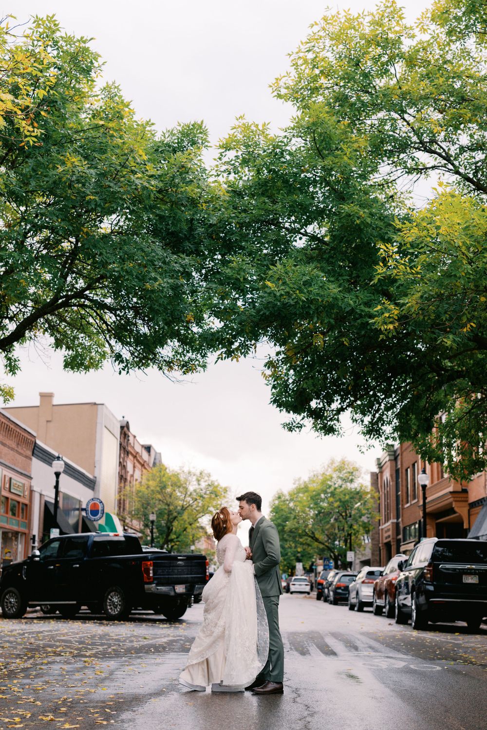 A romantic couple shares a moment on a tree-lined city street during autumn with parked cars and buildings in the background.