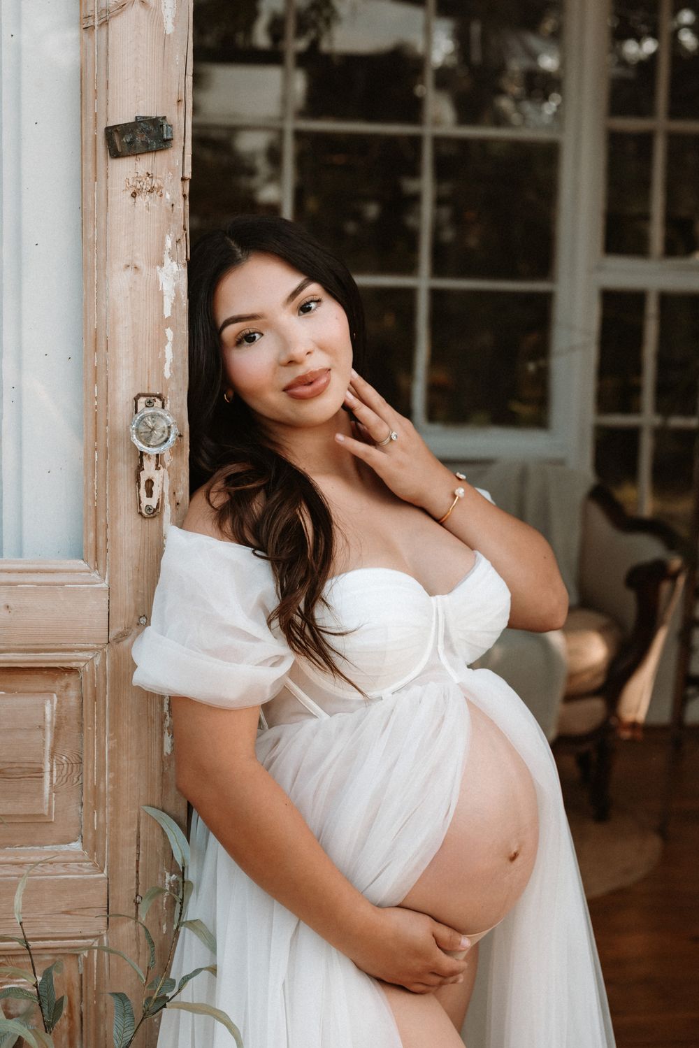 Pregnant woman in white off-shoulder gown cradling baby bump by rustic wooden door.