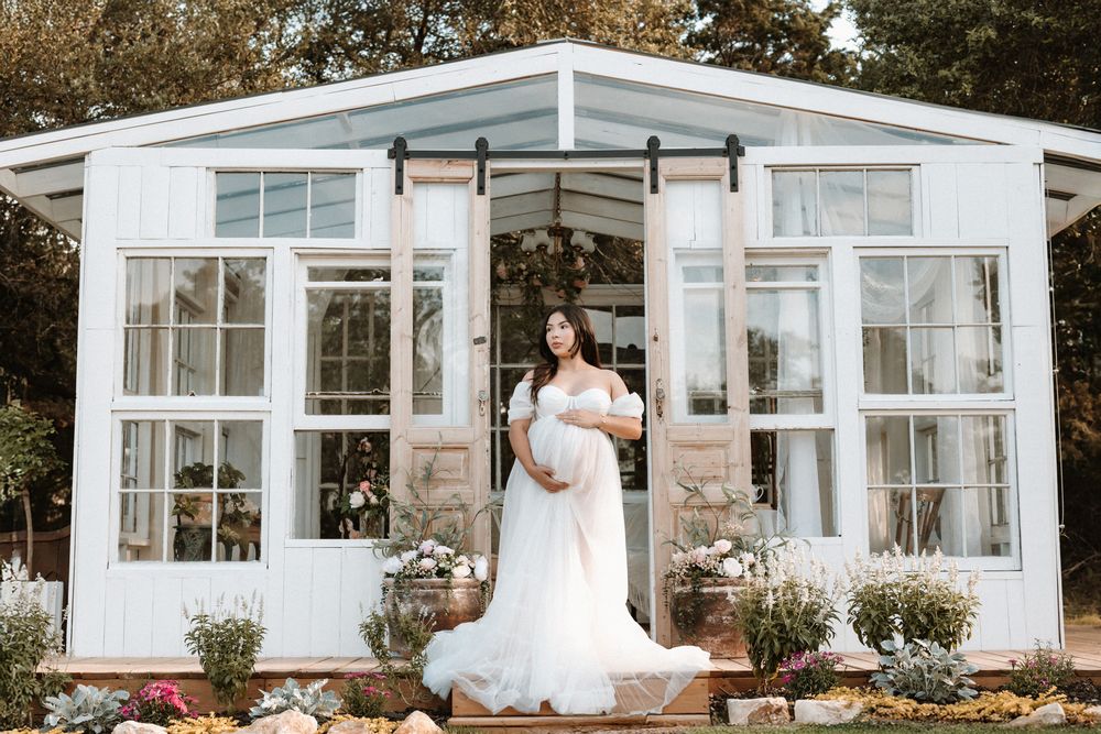 Pregnant woman in flowing white maternity gown posing before a charming white greenhouse.