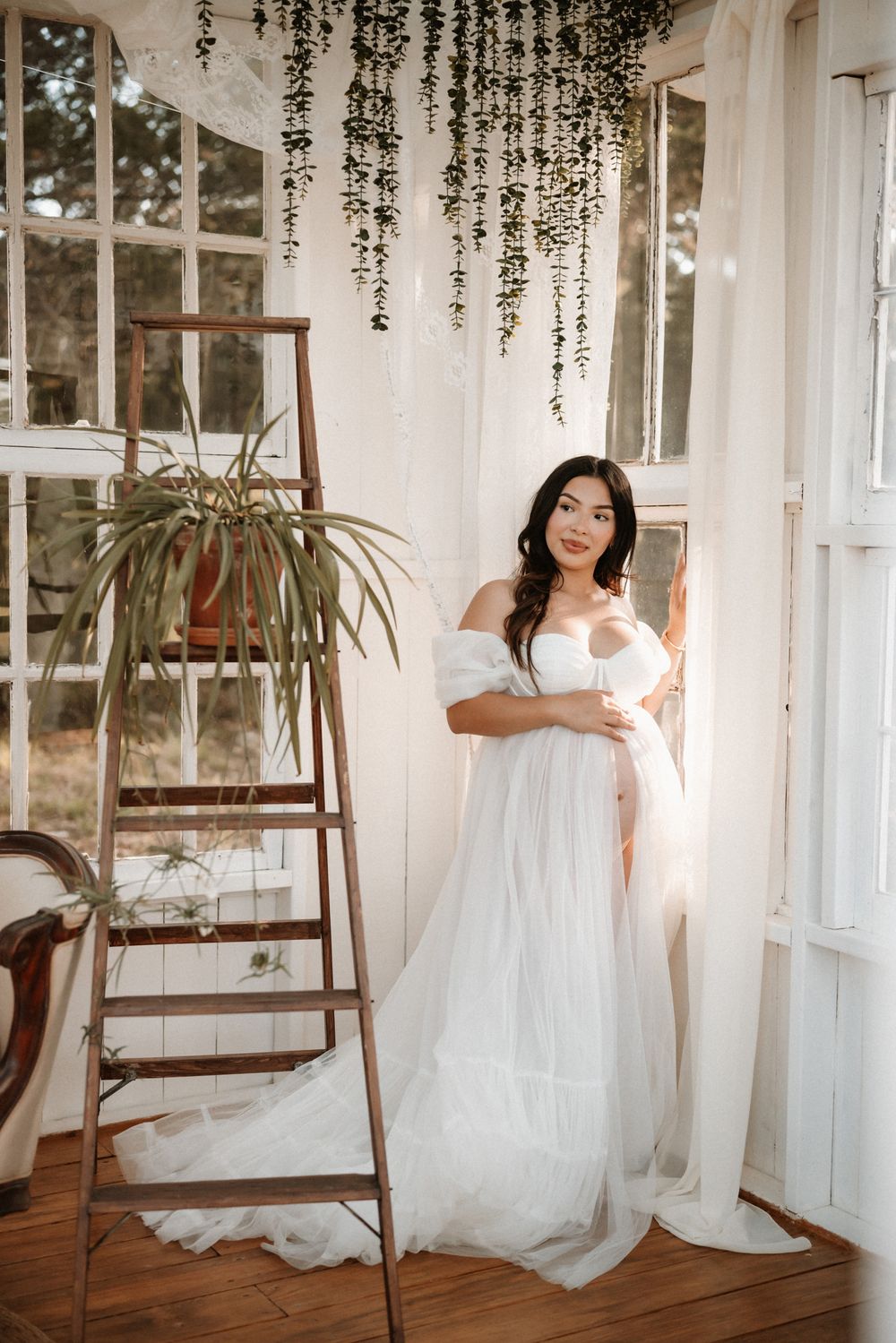 Pregnant woman in white tulle maternity gown poses by sunlit windows with hanging greenery and rustic ladder.
