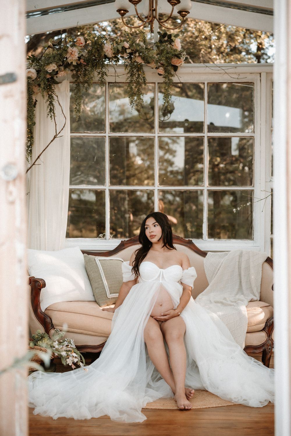 Pregnant woman in white tulle maternity gown sitting on vintage sofa in floral greenhouse studio.