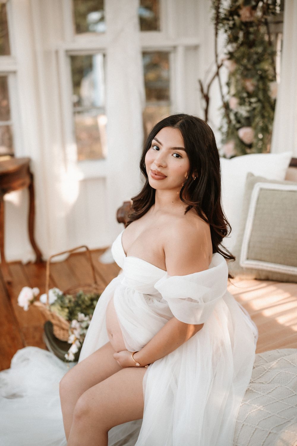Pregnant woman in white off-shoulder tulle gown sitting gracefully in sunlit room with floral decor.