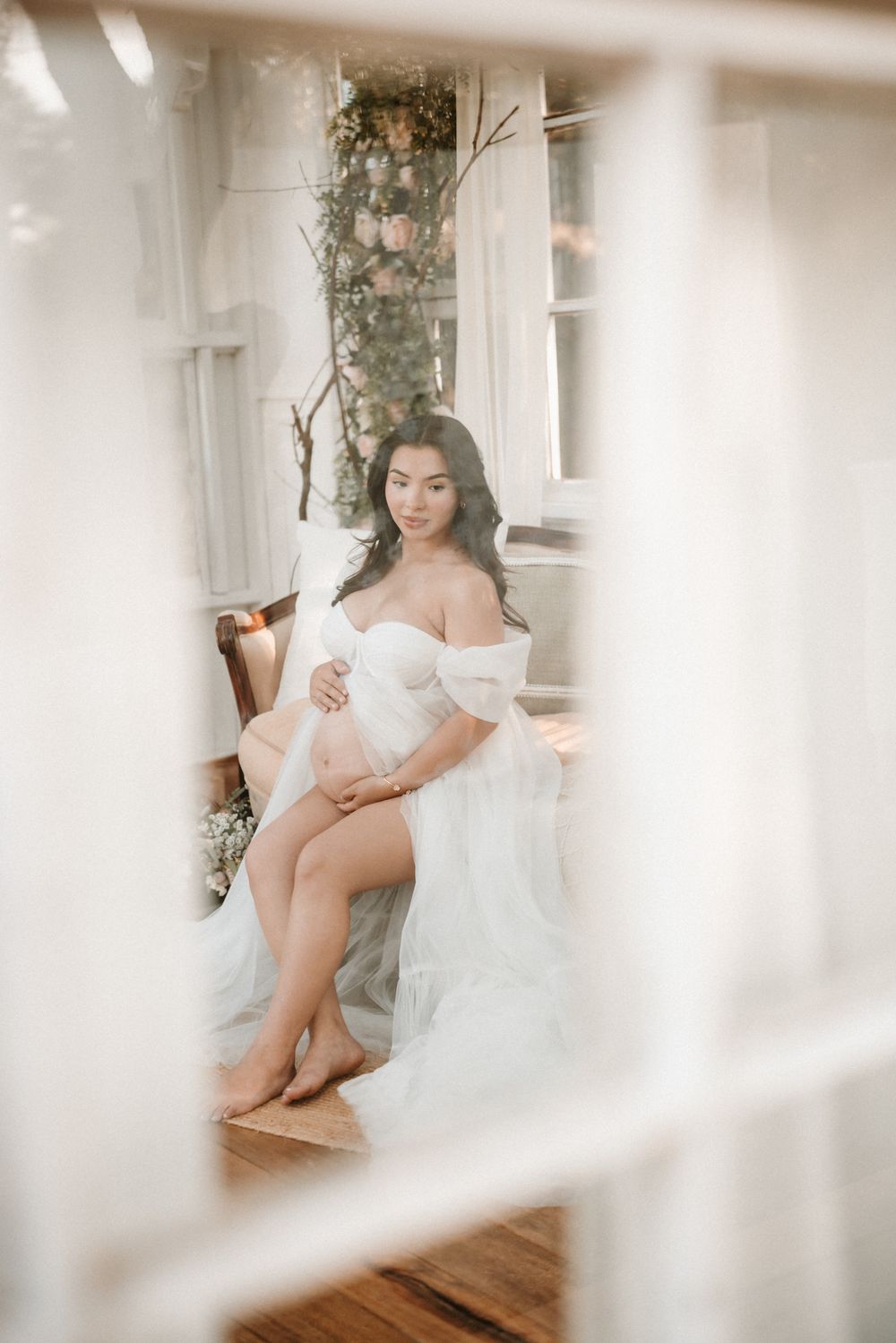Pregnant woman in elegant white tulle off-shoulder gown sits gracefully in chair by window with floral decor.