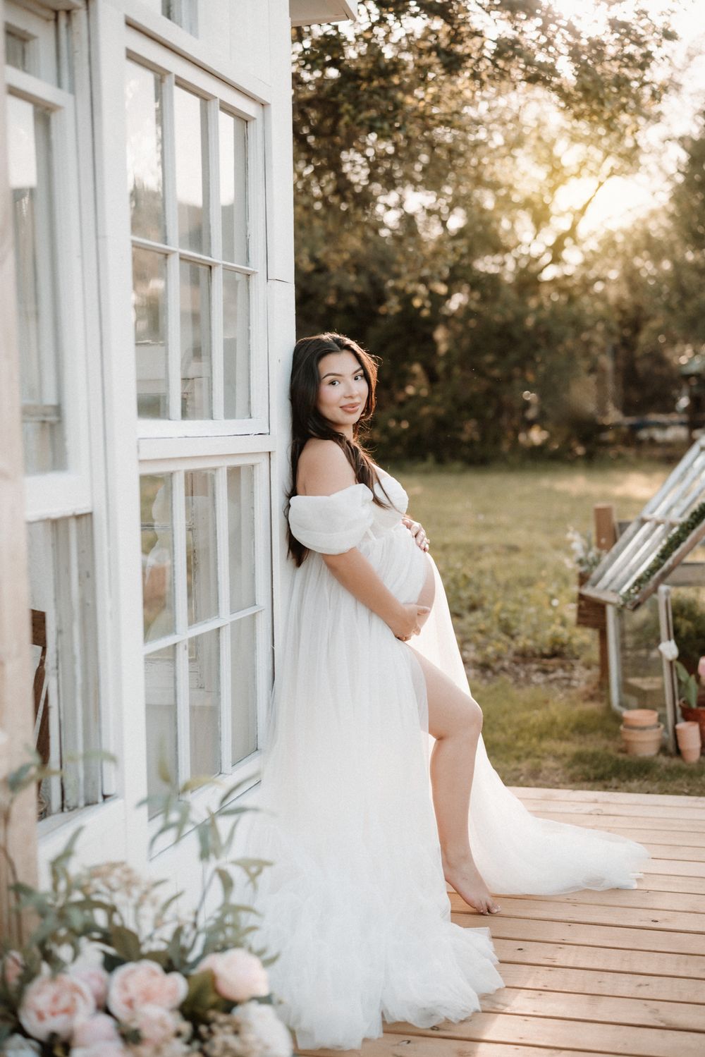 Pregnant woman in white off-shoulder gown posing outdoors at golden hour near white French doors on a wooden deck.