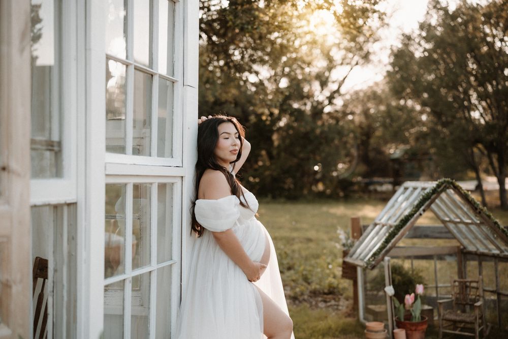 Pregnant woman in white off-shoulder gown leaning against glass door at golden hour outdoors.