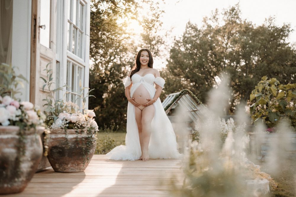 Pregnant woman in white off-shoulder gown poses on sunlit deck surrounded by lush greenery and flowers.