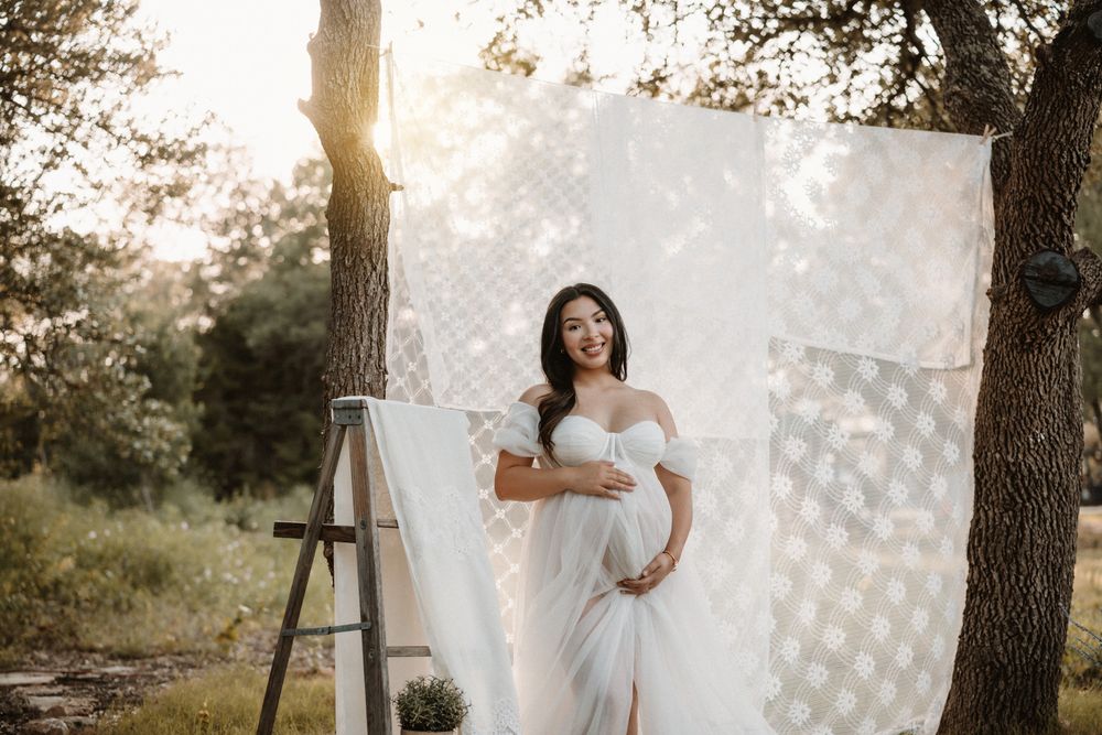 Smiling pregnant woman in white off-shoulder gown stands outdoors in golden sunlight with lace backdrop between trees.