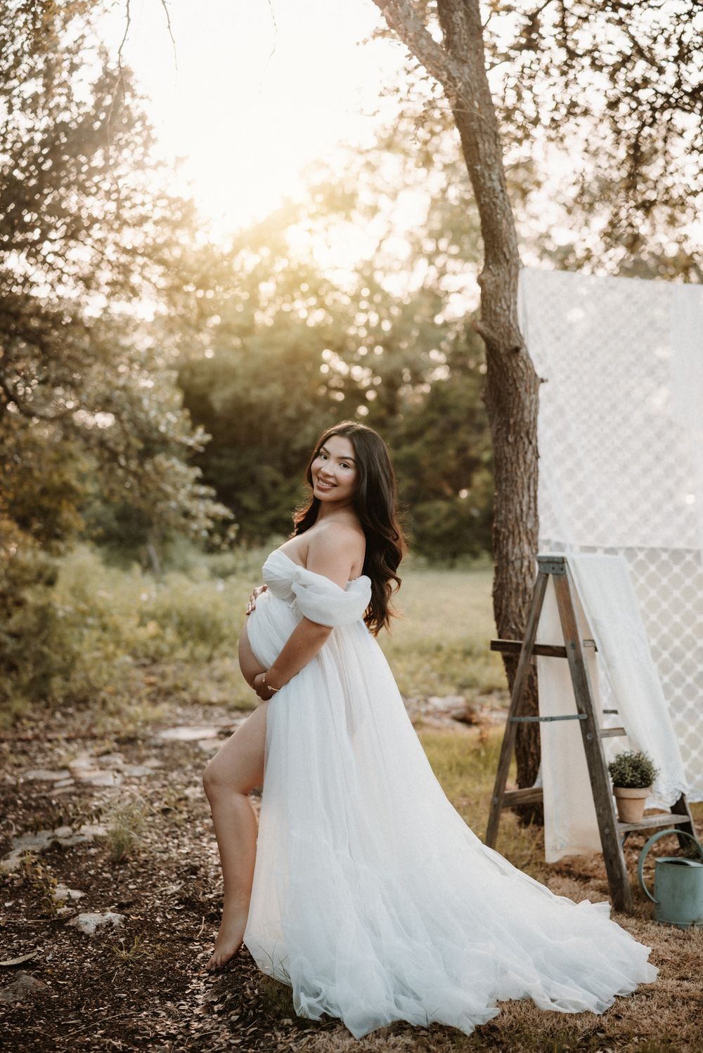 Pregnant woman in white off-shoulder tulle gown posing outdoors at golden hour near trees.