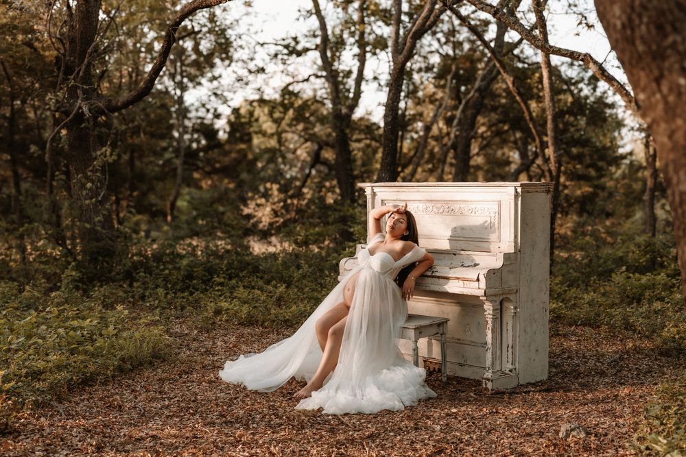 A woman in a flowing white gown poses gracefully beside a vintage white piano in an autumn forest setting.