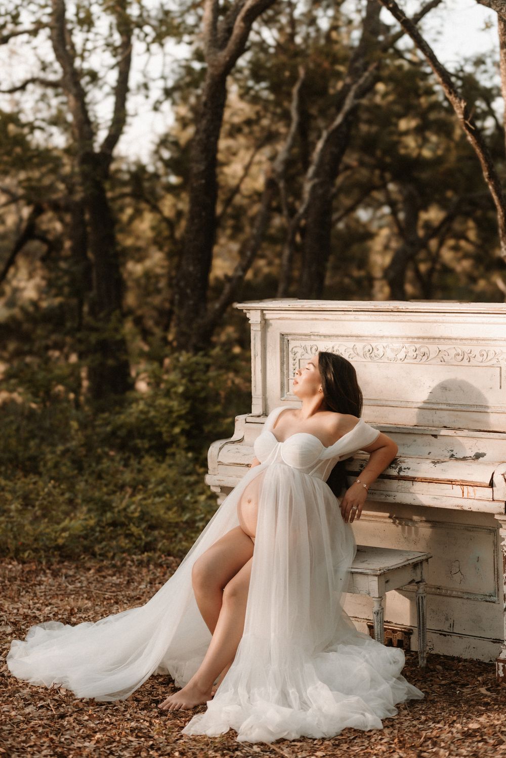Pregnant woman in white tulle gown sits beside vintage white piano outdoors in golden forest light.
