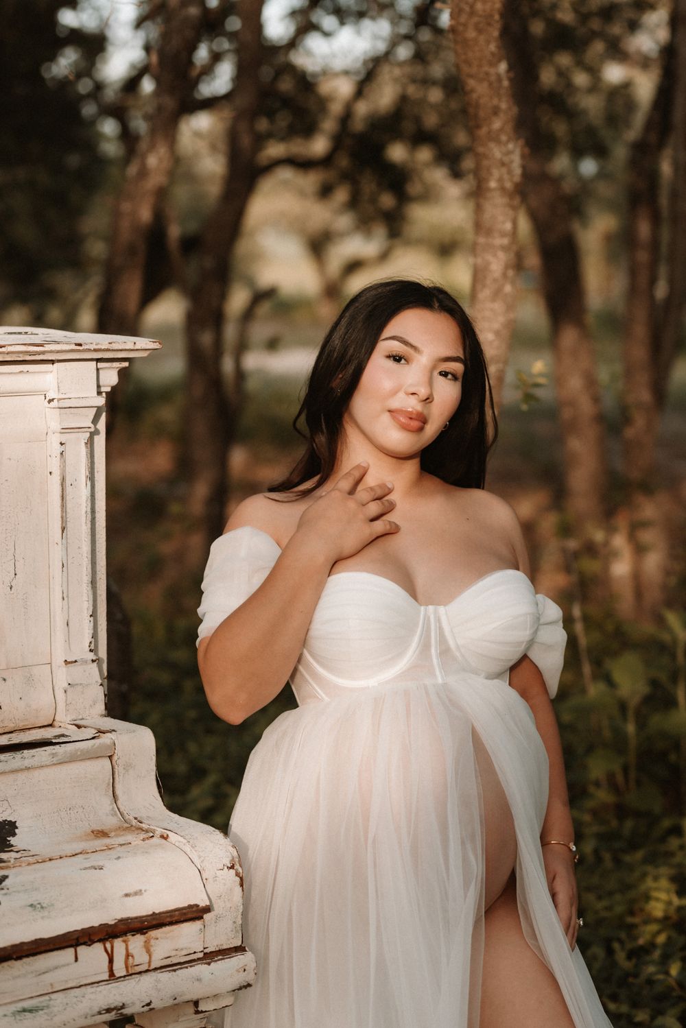 Pregnant woman in white tulle off-shoulder gown poses beside vintage white dresser in outdoor woodland setting.