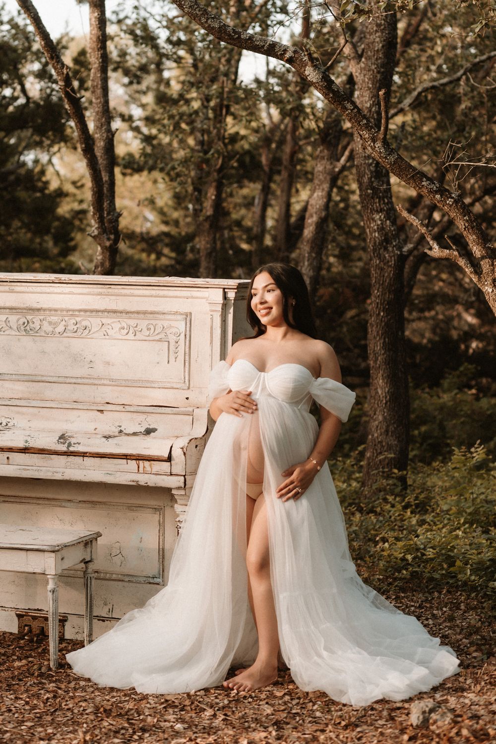 Pregnant woman in flowing white tulle off-shoulder gown poses beside a vintage white piano in a wooded setting.