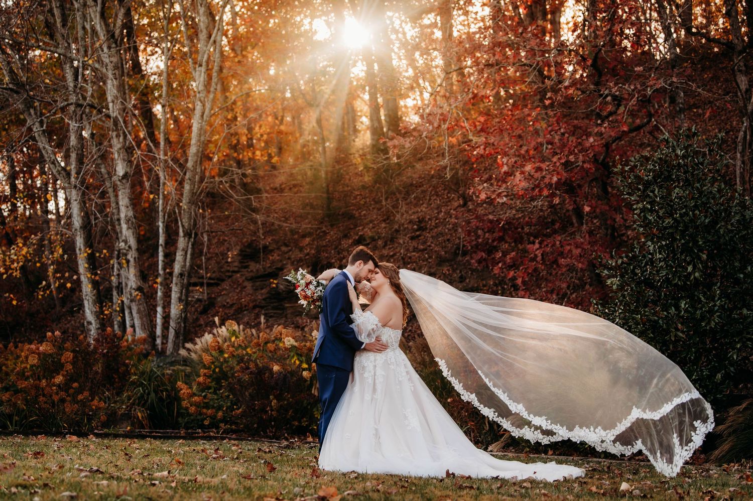 A dramatic wedding portrait with flowing veil against autumn trees at sunset.