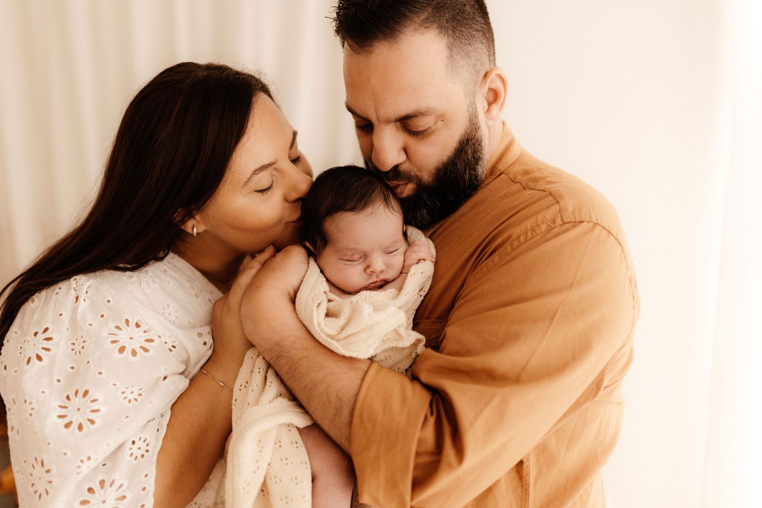 Parents share intimate moment with newborn baby in soft indoor setting.