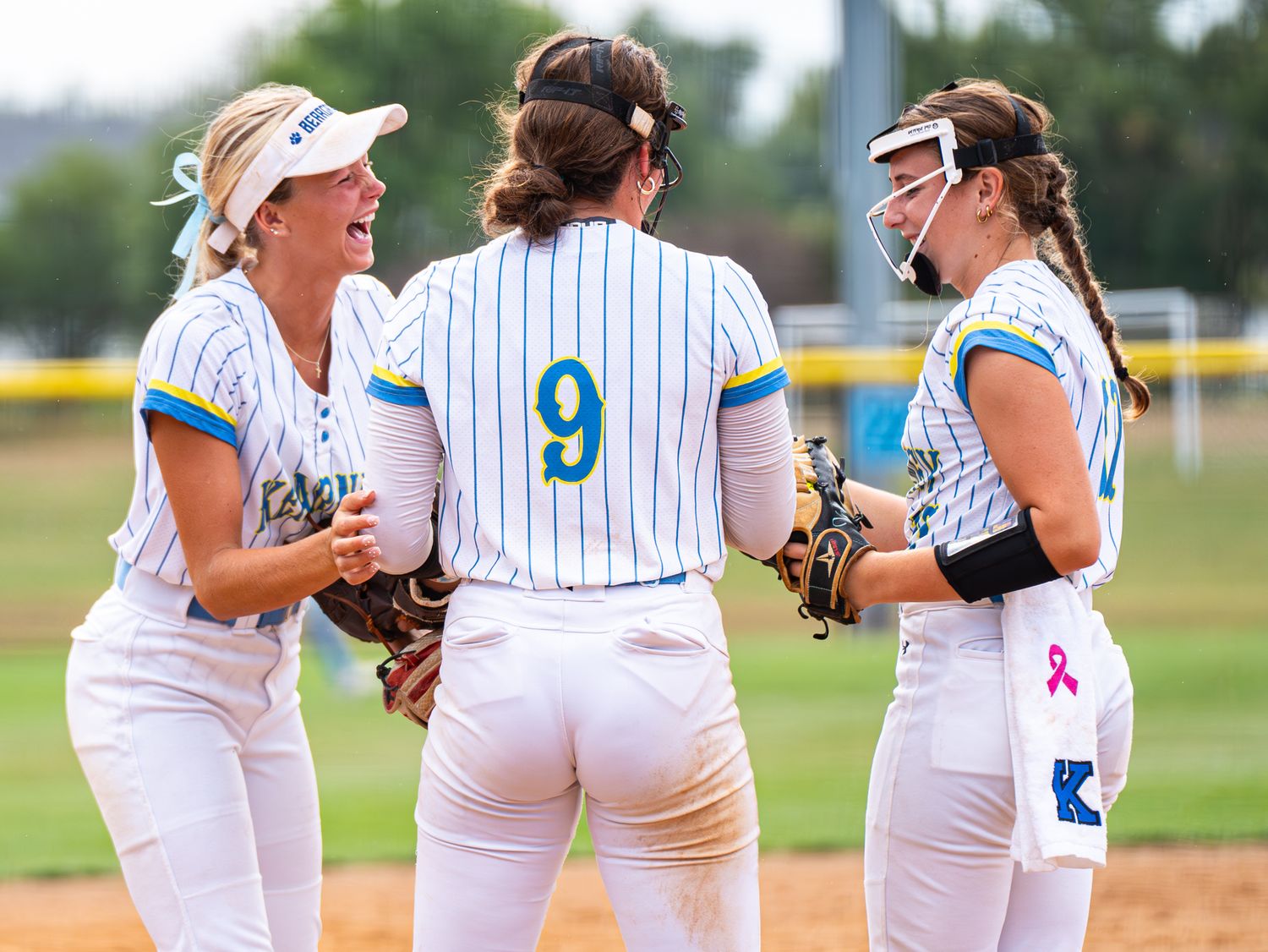 Three softball players in white pinstripe uniforms huddle during game discussion.