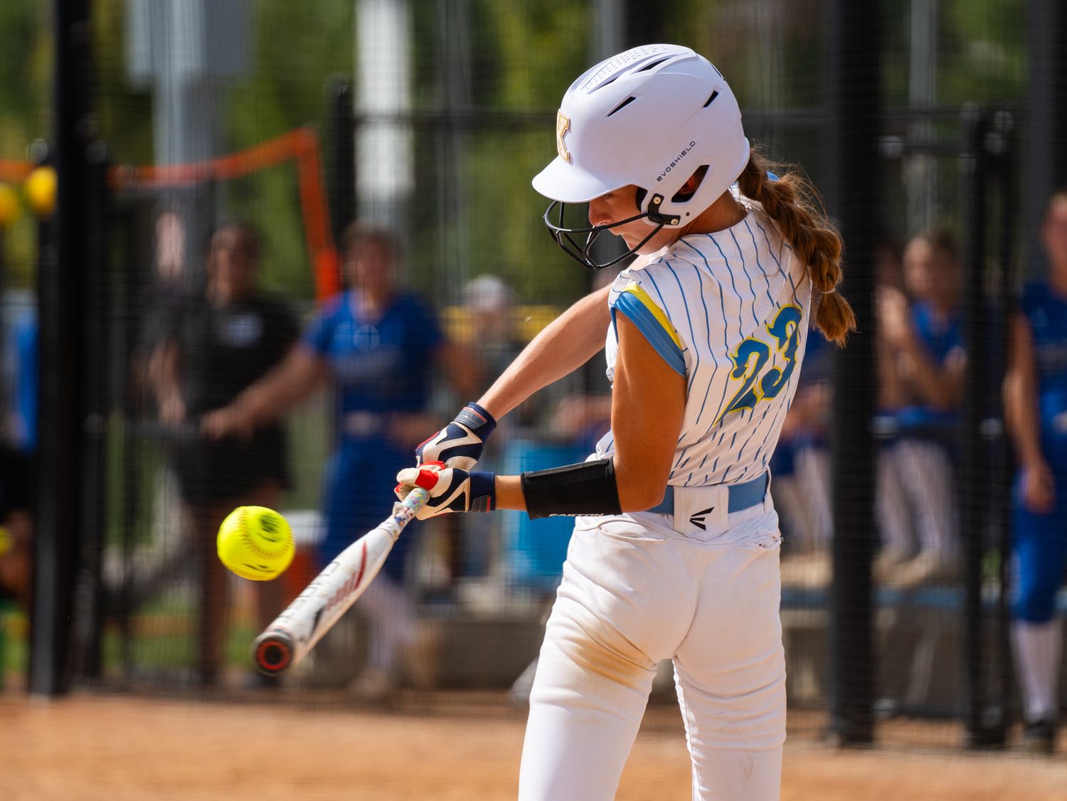 Softball player in white uniform swings bat connecting with yellow ball.