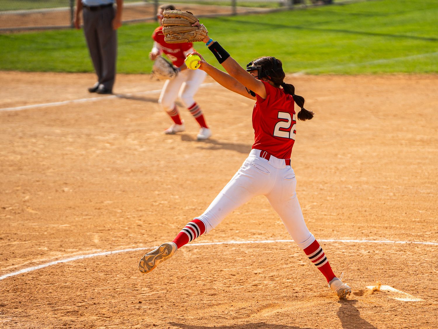 Softball pitcher in red and white uniform delivers pitch from mound.