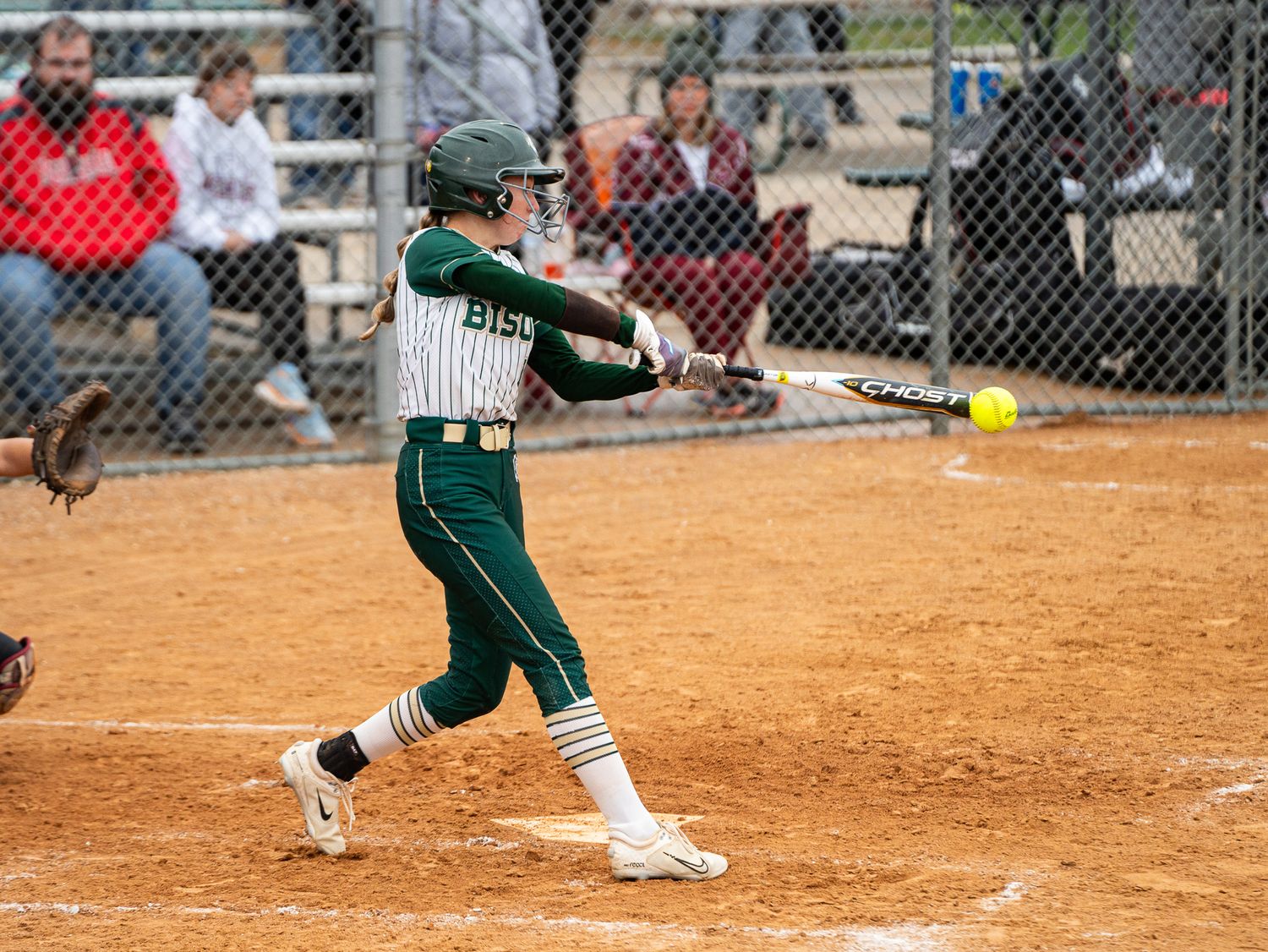 Softball player in green uniform swings bat on dirt infield during competitive game.