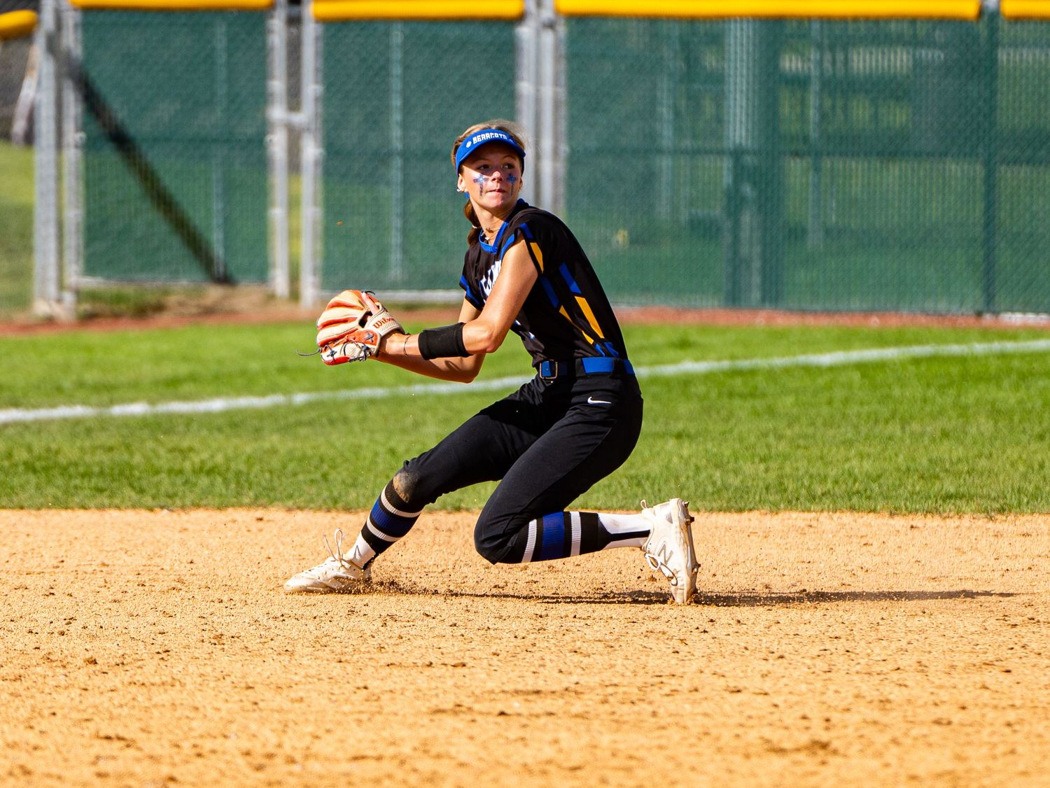 Softball fielder in black uniform crouches ready to catch the ball.