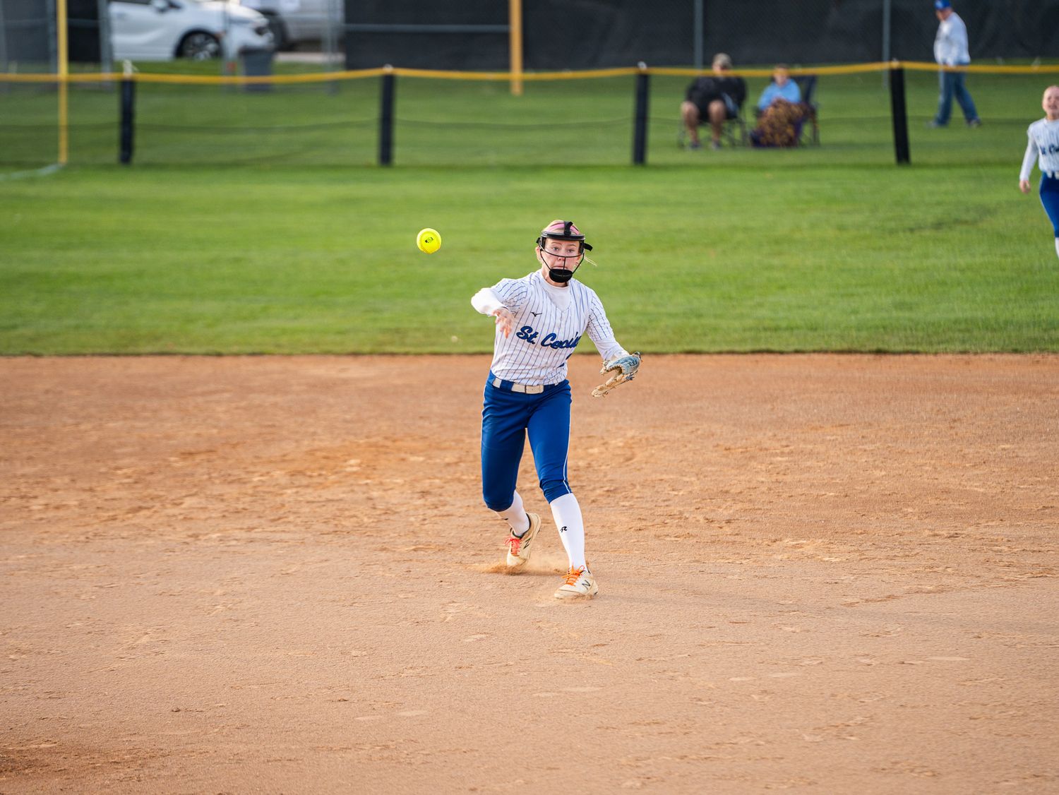 Softball player in blue uniform prepares to throw bright yellow ball on dirt infield.
