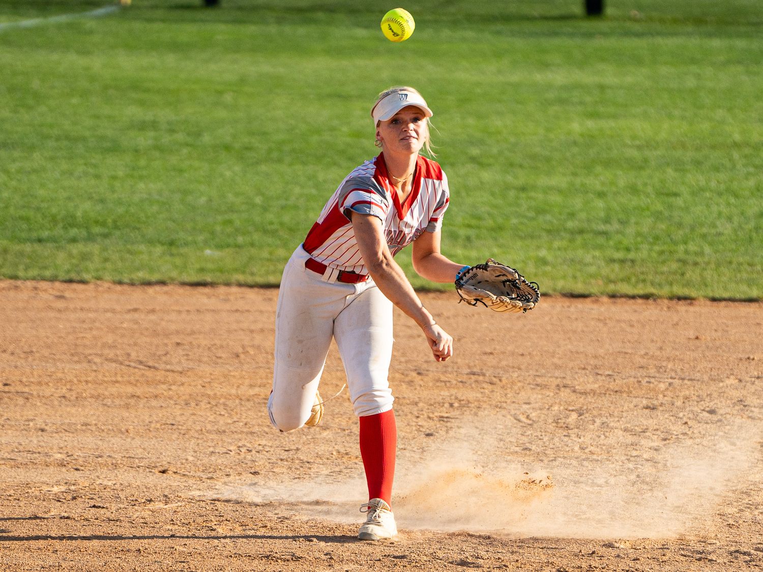 Softball pitcher in red and white uniform delivers pitch with dust kicking up.