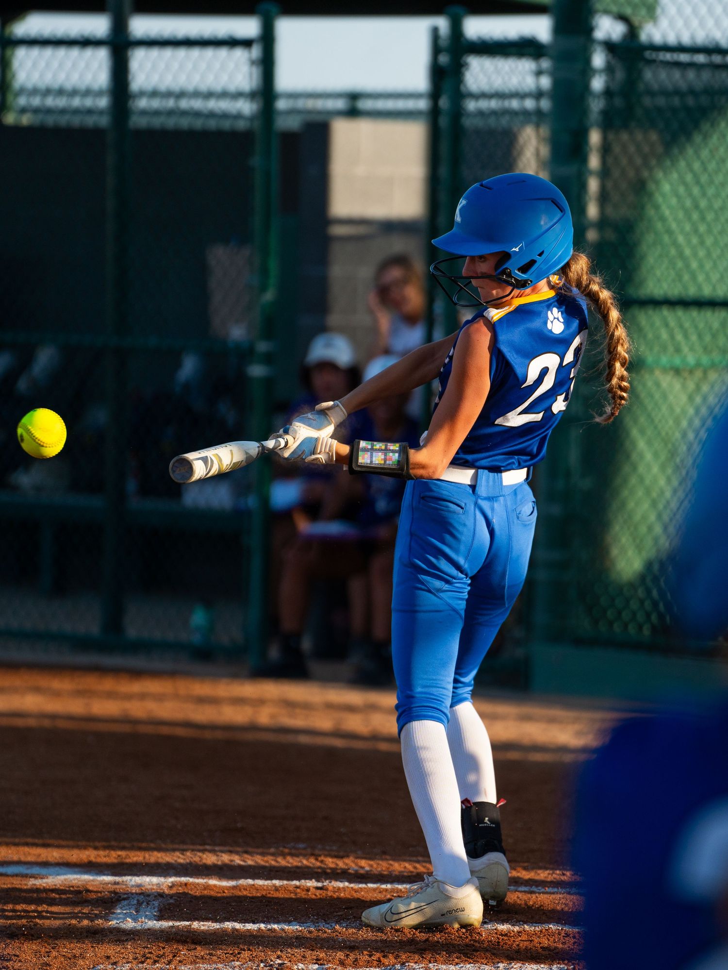 Softball batter in bright blue uniform swings at pitch during evening game with fence visible in background.