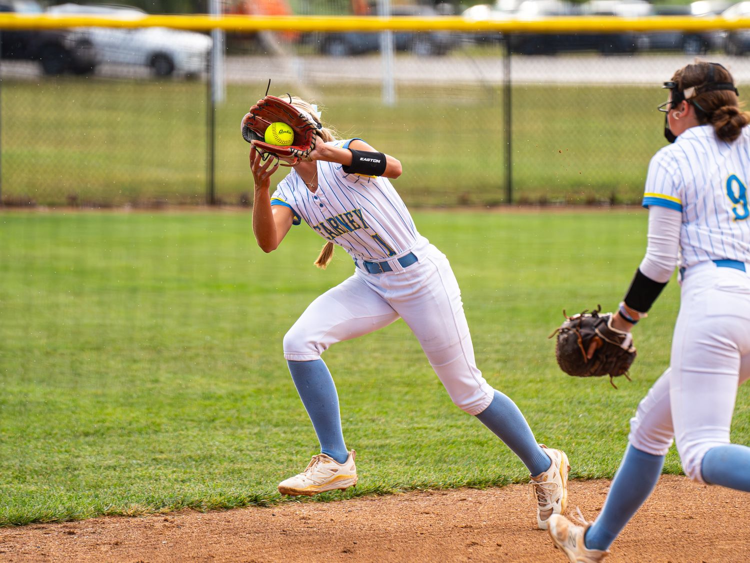 Softball player in white uniform and blue socks reaches to catch the ball on a grass field during a game.