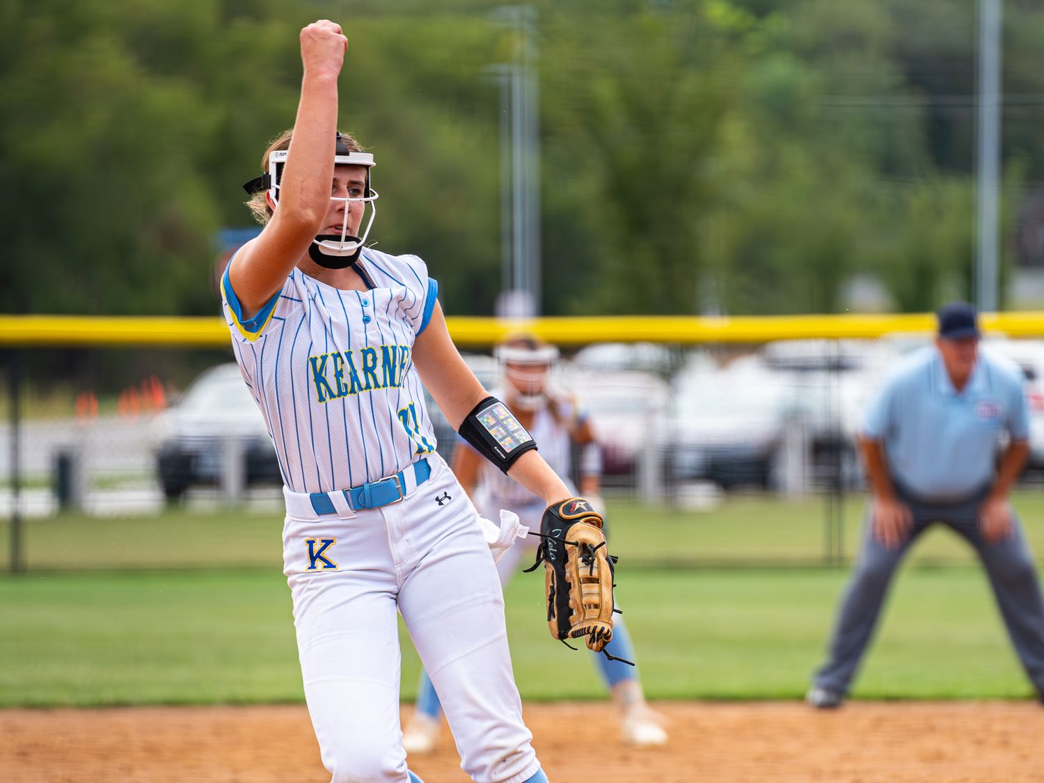 Softball player in light colored uniform winds up for a pitch on the field with umpire watching.