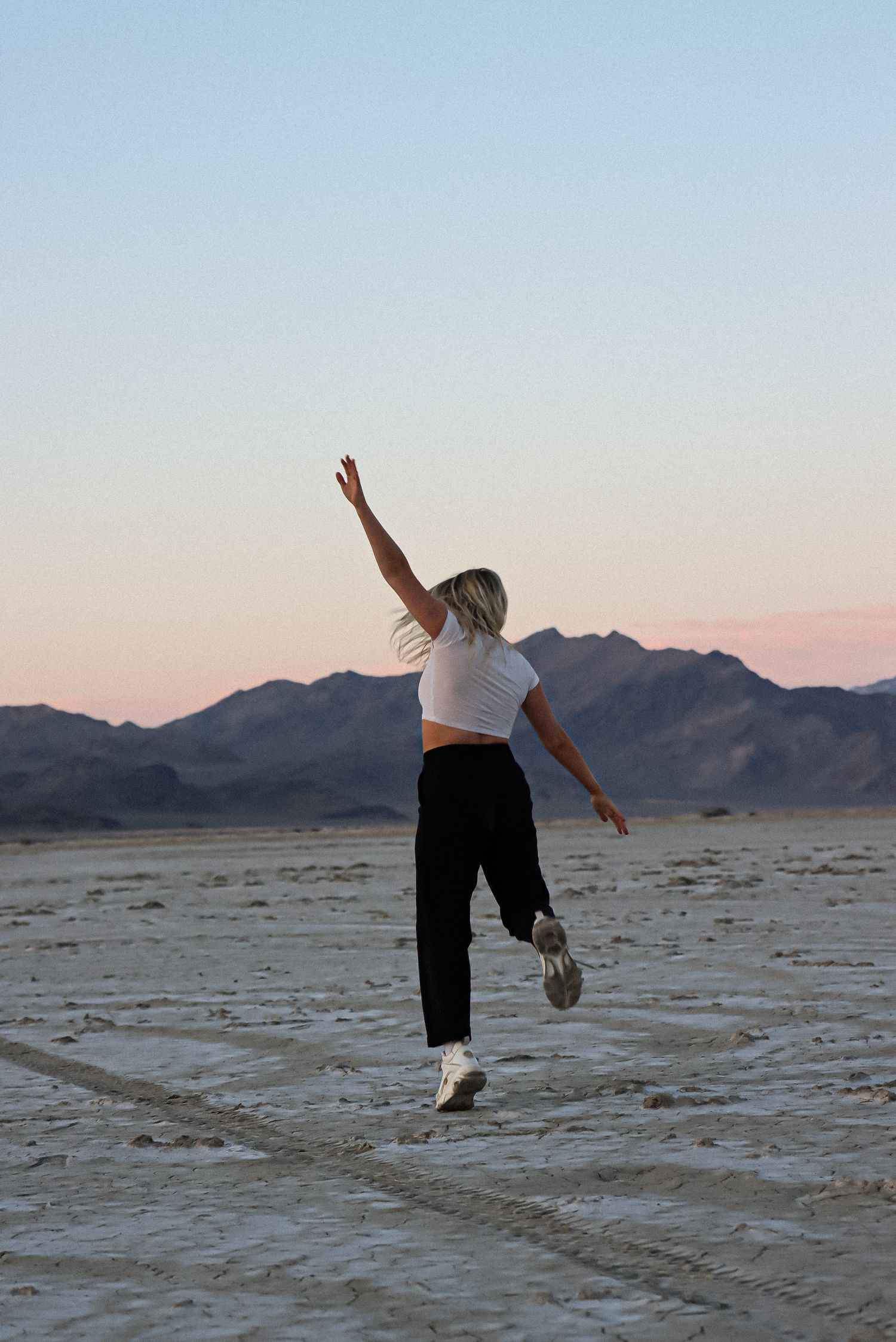 Cora Emilie Media -A person jumping with outstretched arms on a desert plain at sunset with mountains in the background.