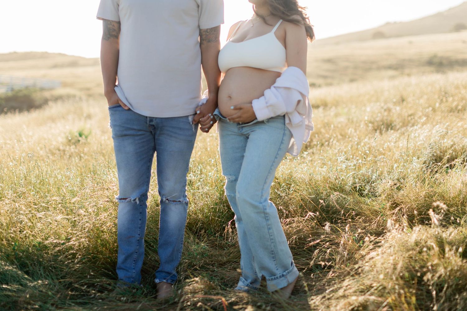 Couple holding hands in golden field showing pregnant belly in casual denim outfits.