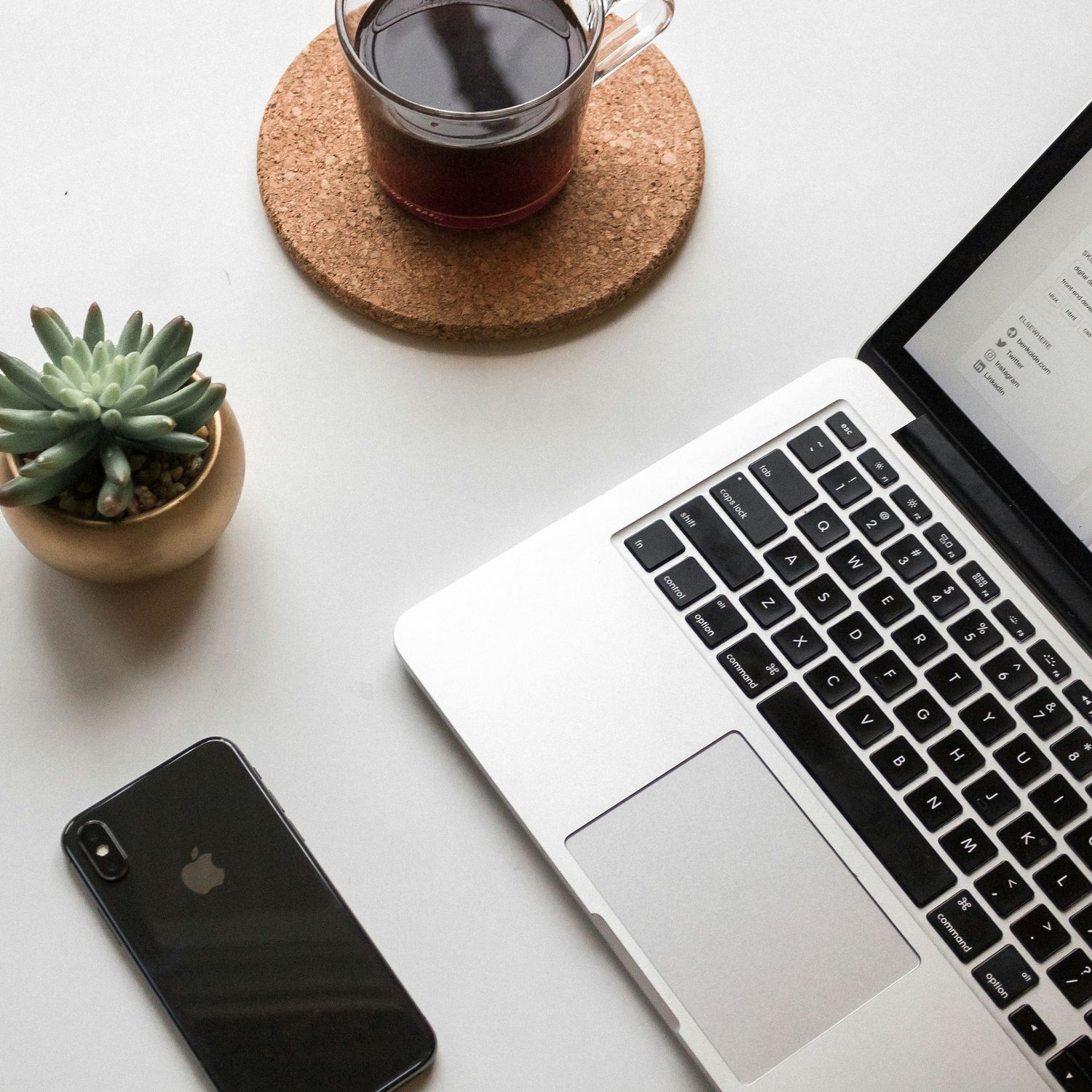 Minimalist workspace setup with MacBook, iPhone, coffee cup and succulent plant on white desk.