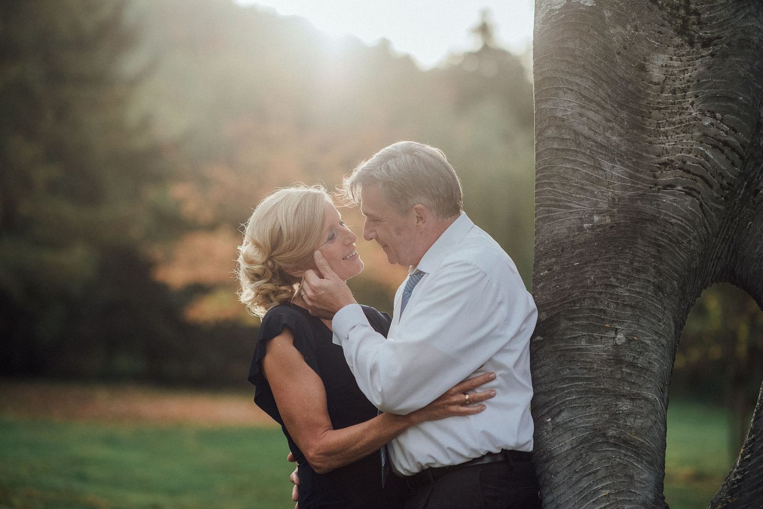 A romantic couple embrace and kiss by a tree trunk during golden hour sunlight in a park setting.