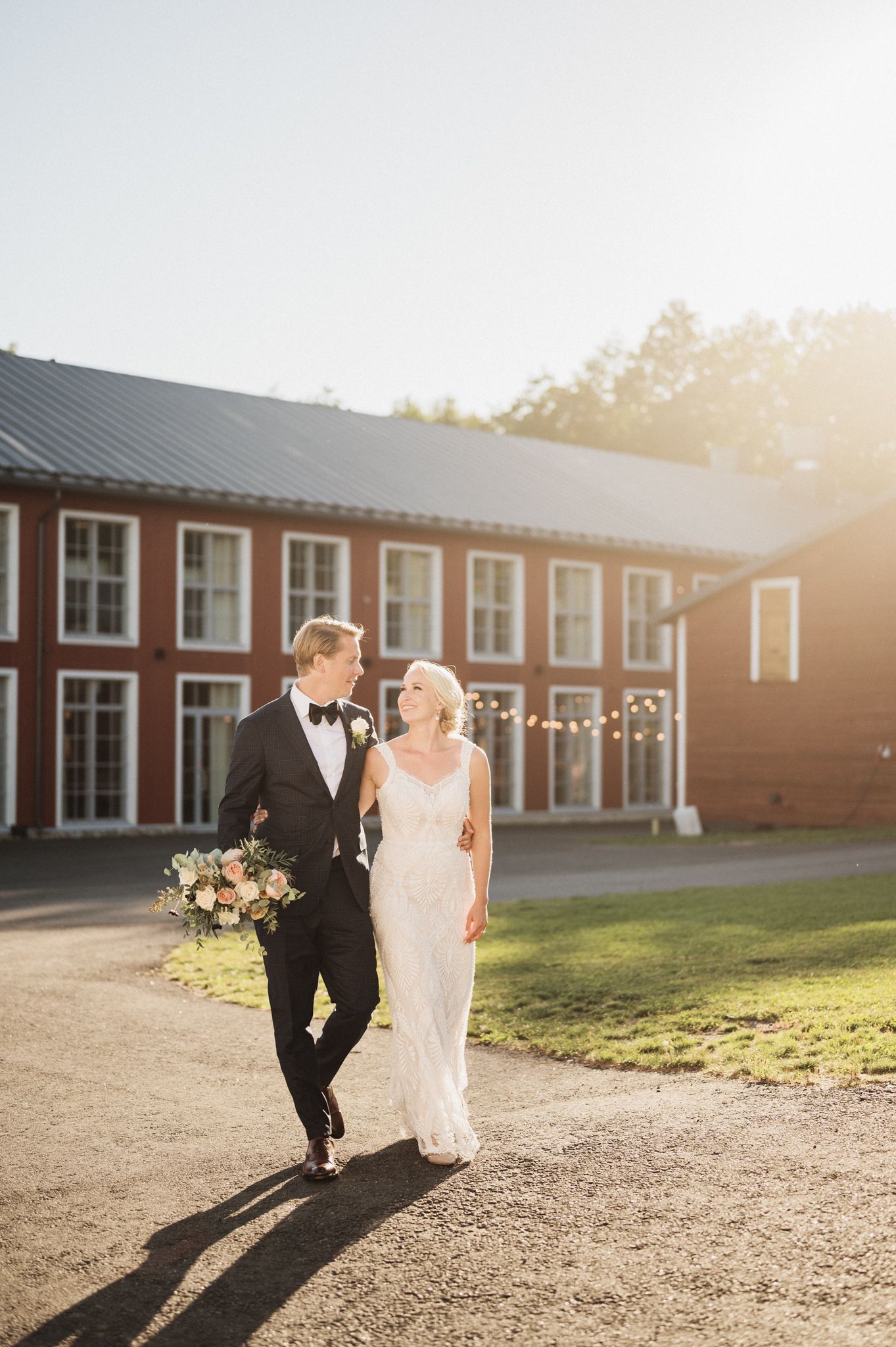 A couple in wedding attire walks together at golden hour outside a rustic red barn building.