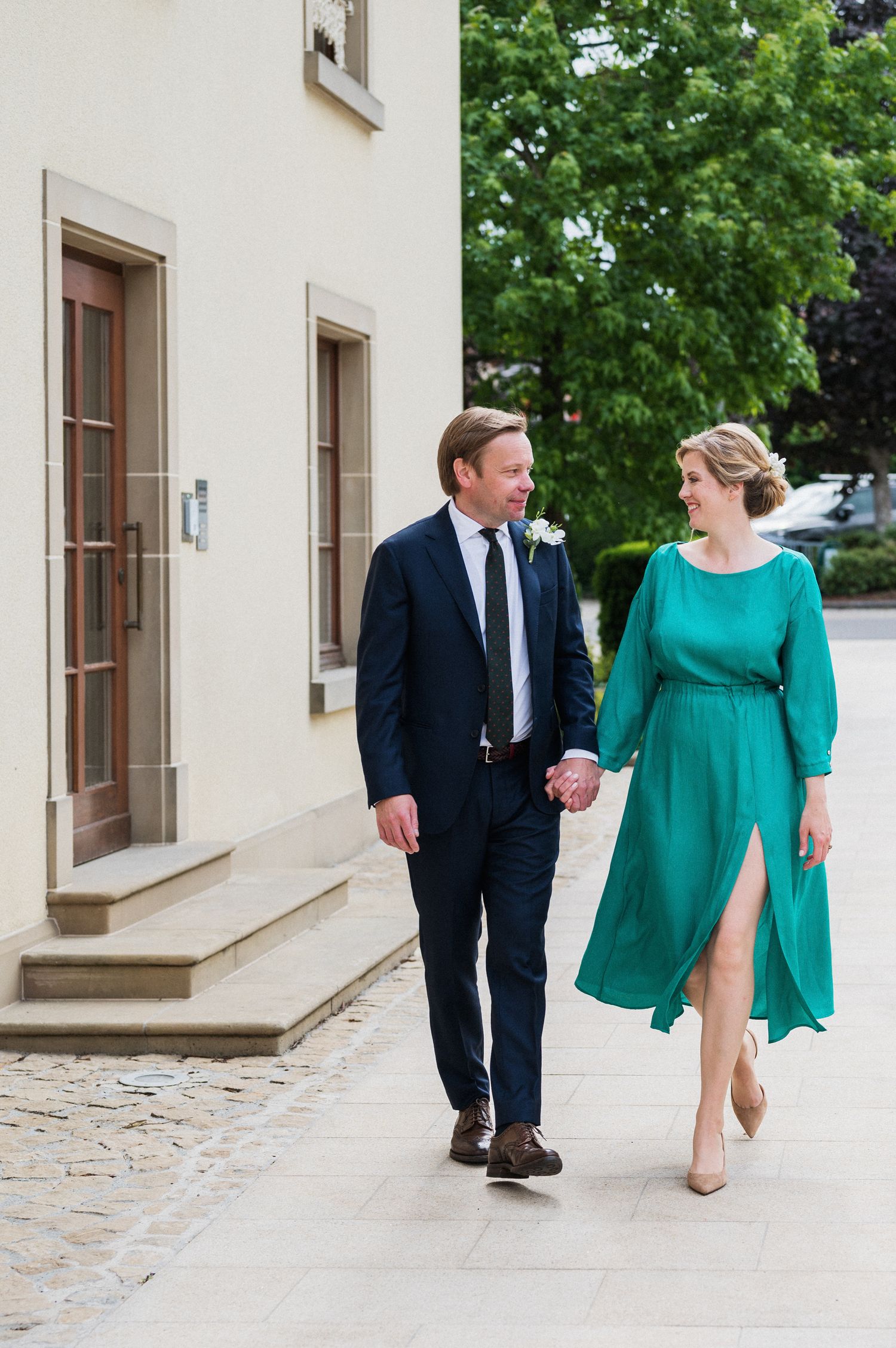 A couple walks hand in hand outdoors, with the person wearing a navy suit and the other in a flowing emerald green dress.