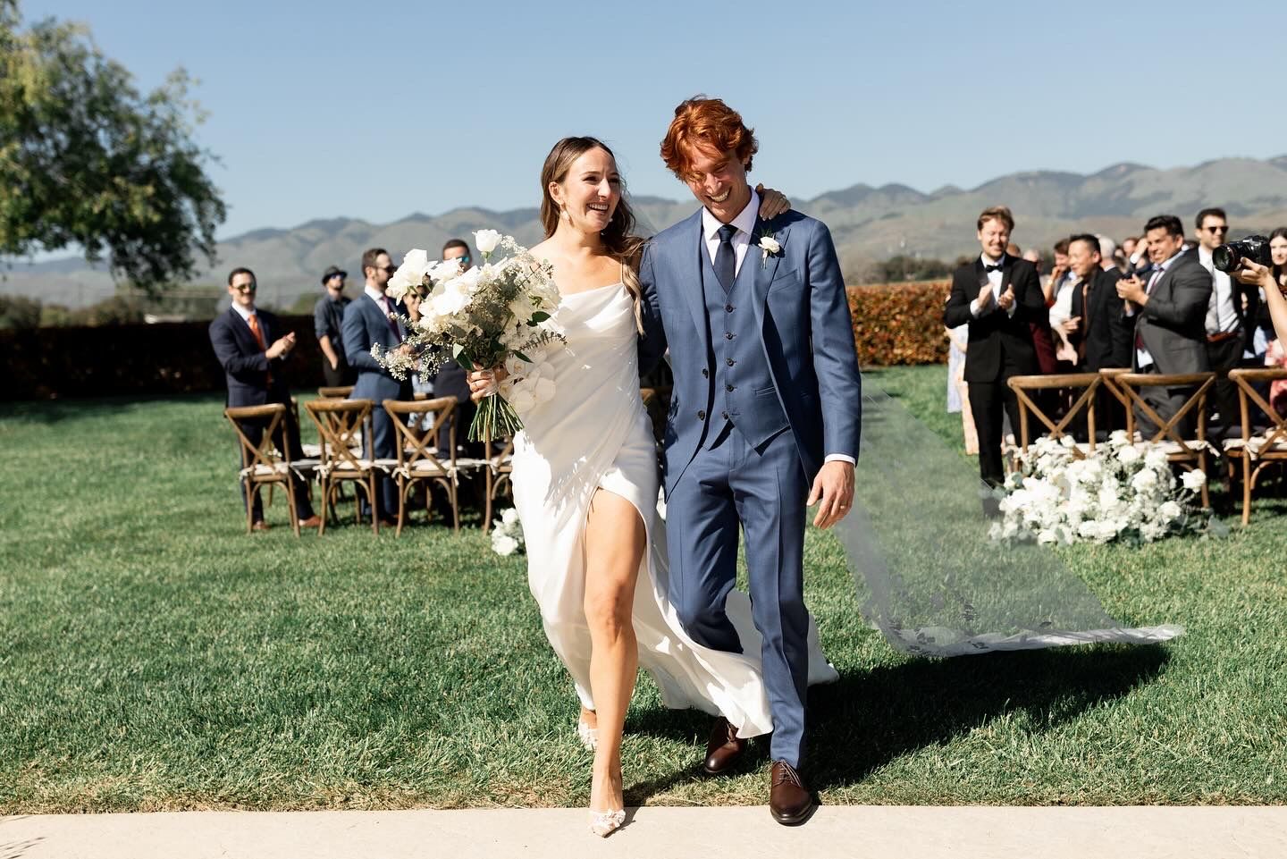 A couple exiting their romantic outdoor wedding ceremony setup with wooden chairs and mountain views in the background.