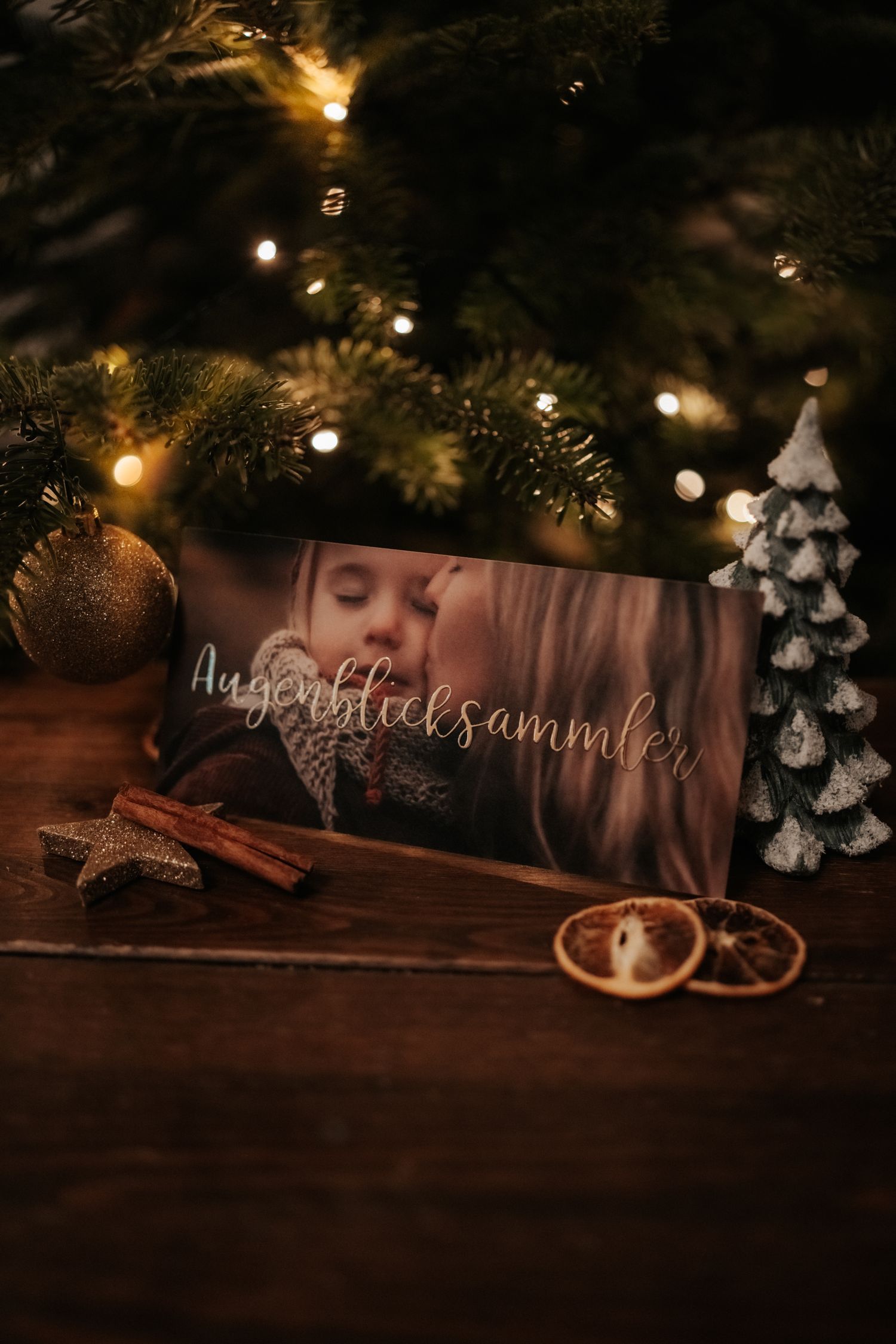 Cozy Christmas scene with decorative card, cinnamon sticks, dried orange slices and glowing tree lights in the background.