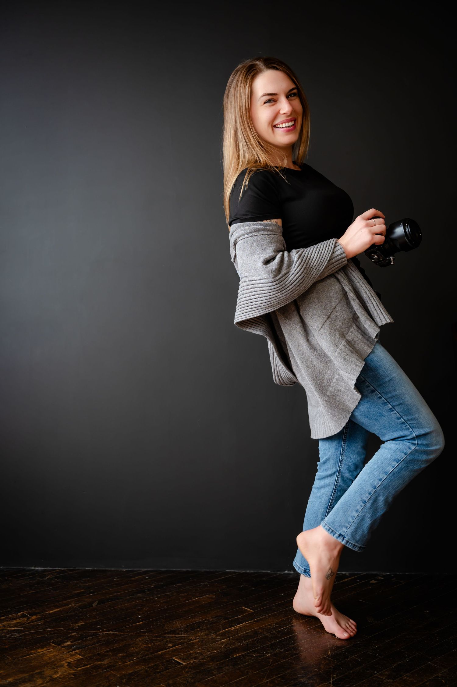 Tattooed photographer in rust-colored top and blue jeans sits with camera against dark background Lauren Perrin Photo