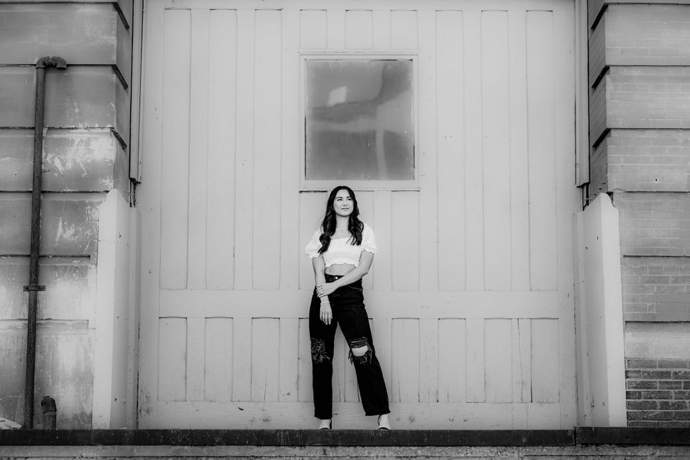 Artistic black and white photograph of a figure standing against a white industrial wall with a window above.