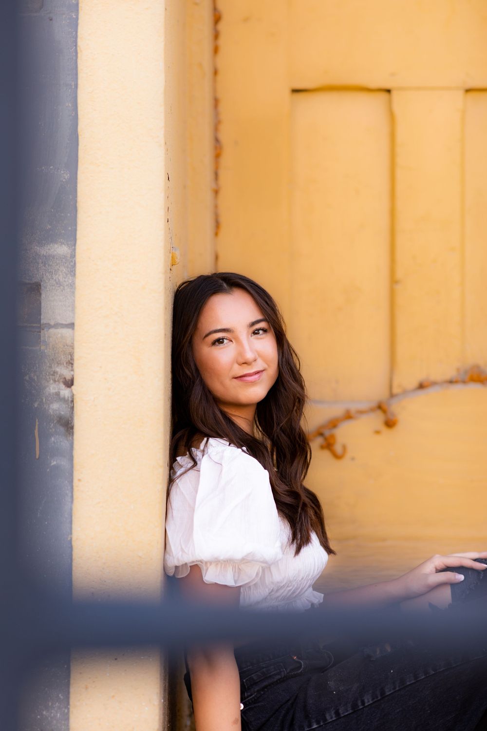 Person wearing a white top poses thoughtfully against a yellow wall in warm natural lighting.