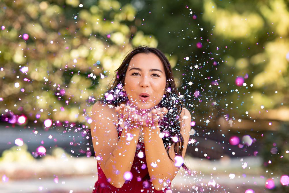 Someone in a red top blows glitter confetti in an outdoor setting with sunlit bokeh background in summer.