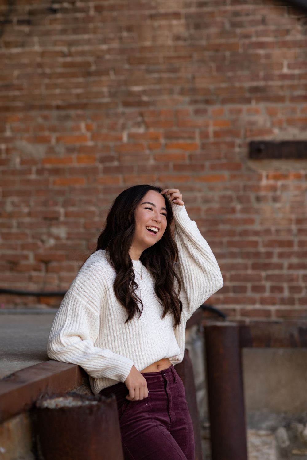 Laughing person in cozy cream sweater and burgundy pants poses against rustic brick wall backdrop.