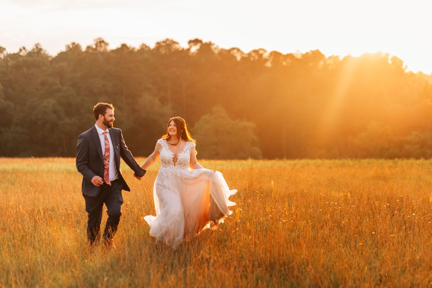 A couple runs hand in hand through a golden field at sunset.