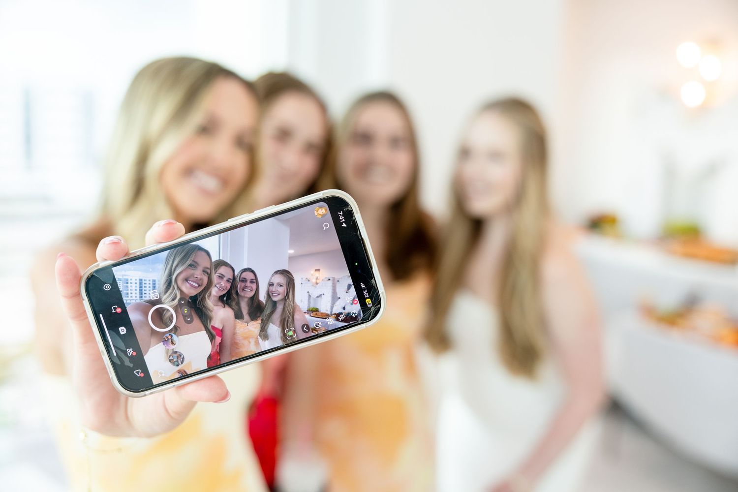 Group of four young women taking a selfie with a smartphone at a bright indoor gathering.