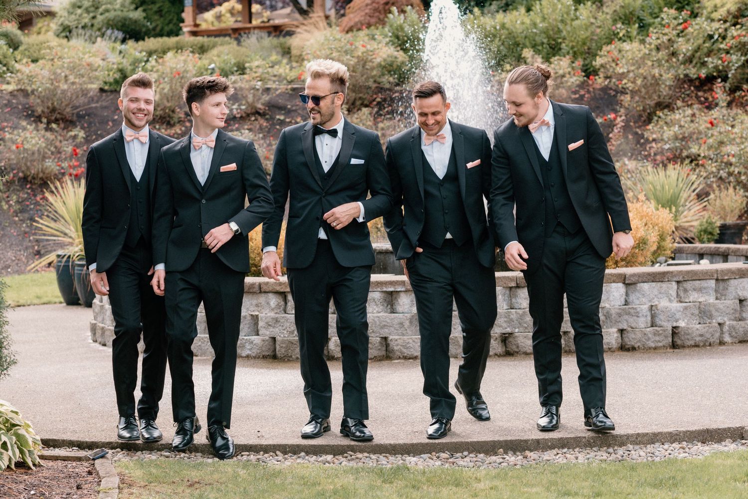 Groomsmen in black tuxedos walk together outdoors during wedding celebration.