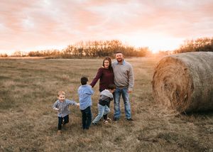 Family of four posing near a hay bale in a golden field at sunset.