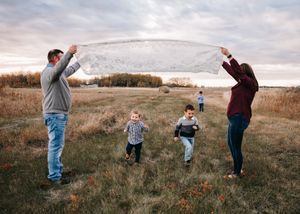 Parents lifting a white blanket over two young boys running underneath in an autumn field.