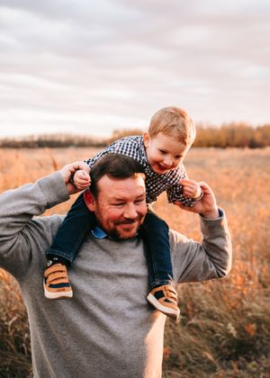 Smiling father carrying a happy toddler on his shoulders in a golden autumn field at sunset.