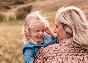 A smiling toddler girl touches her blonde mothers face in a warm outdoor portrait.