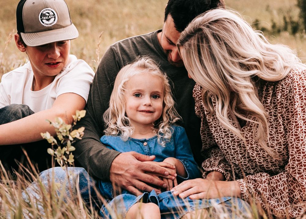 A family of three sits together in a golden field with parents gazing at their daughter.