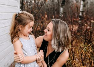 A mother and young daughter share a joyful laugh together against a white wall outdoors.
